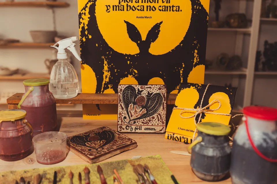El socarrat «Oh foll amor» damunt del banc del taller, amb el cartell groc dels versos d'Ausiàs March darrere i una peça ja embolicada.